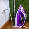 A purple and white iron is plugged into a white smart plug in a wall socket. The background is a mix of green artificial grass and a wooden surface. Produktbild-Vorschau