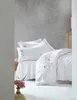 A neatly made bed with white bedding and pink floral embroidery, set against a textured gray wall. Podgląd zdjęcia produktu