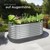 A raised garden bed filled with plants on a patio, with a garden and trees in the background. Produktbild-Vorschau