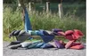A collection of colorful cushions and blankets are arranged on a wooden bench and on the ground in an outdoor setting with a body of water and reeds in the background. Produktbild-Vorschau