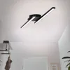 A modern black LED ceiling light fixture with two angled arms, mounted on a white ceiling in a hallway with a coat rack and a door. Produktbild-Vorschau