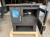 A black wood-burning stove is shown in a workshop or storage area, resting on a wooden pallet. Produktbild-Vorschau