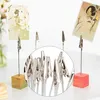 A close-up view of several metal alligator clips attached to thin wires, each mounted on a small wooden block base. Two small artistic cards are displayed on clips in the background. Produktbild-Vorschau