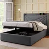 A grey upholstered bed frame with the mattress lifted to reveal storage space underneath, in a room with a window and wall paneling. Produktbild-Vorschau