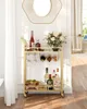 A gold bar cart filled with bottles, glasses, and fruit, placed in a room with white walls and curtains. Produktbild-Vorschau
