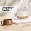 Two cappuccino cups are displayed on white pedestals with wooden saucers, set against a softly lit background with abstract shadows. Produktbild-Vorschau