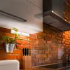 A kitchen counter with a brick wall background, featuring under-cabinet lighting, a potted plant, and a knife block. Produktbild-Vorschau