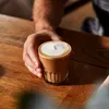 A hand holding a cup of coffee with foam on a wooden table. Produktbild-Vorschau
