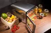 A stainless steel food dehydrator with its door open, displaying trays of dried fruit, surrounded by fresh fruits, vegetables, and jars of dried produce. Podgląd zdjęcia produktu