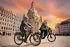 Two people riding electric bikes in a European city square with a large church in the background. Produktbild-Vorschau