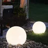 Two illuminated spherical garden lights are placed on a lawn and a stone-covered area next to a patio with outdoor furniture. Produktbild-Vorschau