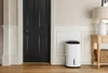 A white dehumidifier is placed next to a dark gray door in a hallway, with a wooden table and lamp in the background. Podgląd zdjęcia produktu
