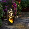 A solar-powered garden lantern with a tree design is placed on a stone pathway, surrounded by plants and flowers. Produktbild-Vorschau