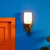 A wall light with a camera is mounted on a blue wall next to an orange door. Produktbild-Vorschau