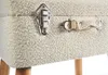 A close-up view of the textured fabric surface of a white storage ottoman or trunk, showing two metal latches and a fabric handle. Náhled obrázku produktu