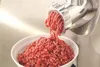 A meat grinder is shown in operation, mincing meat into a white bowl. Podgląd zdjęcia produktu