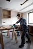 A man is using a circular saw to cut a piece of wood on sawhorses in what appears to be a room under renovation. Produktbild-Vorschau