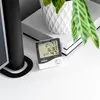 A digital thermometer and hygrometer sits on a white surface next to a black speaker and a white gaming console, with a potted plant in the background. Anteprima dell’immagine prodotto