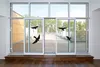 Black bird decals are applied to a large sliding glass door, creating the illusion of birds in flight. The scene is set indoors, looking out onto a wooden deck with trees in the background. Produktbild-Vorschau