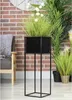A black metal plant stand with a green plant in a black planter, placed on a wooden floor in front of a brick wall and a chair. Produktbild-Vorschau