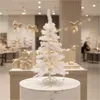 A white artificial Christmas tree decorated with lights and ornaments, placed on a marble-top pedestal in a store setting with gift boxes in the background. Produktbild-Vorschau