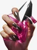 A hand with long, metallic silver nails holds a pink, faceted perfume bottle. Podgląd zdjęcia produktu