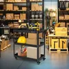 A black three-tier tool cart filled with various tools and boxes, placed in a warehouse setting with shelves of boxes in the background. Náhled obrázku produktu