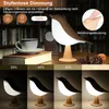 A bird-shaped lamp on a table with books and a cup, demonstrating different brightness levels. Produktbild-Vorschau