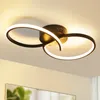 A modern black ceiling light with two intertwined rings, illuminated with warm white light, mounted on a white ceiling. Voir les images du produit