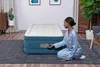 A woman in pajamas is kneeling next to an inflatable air mattress, operating its built-in pump. Produktbild-Vorschau