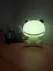 A frog-shaped LED night light is placed on a wooden desk, with some stationery items in the background. Náhľad obrázku produktu