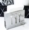 A stainless steel napkin holder with cutouts of a fork, knife, and spoon, filled with white napkins, sits on a marble-like surface. In the background, a dark woven basket with a geometric pattern is visible. Produktbild-Vorschau