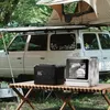 A portable dishwasher and power station are set up on a wooden table in a campsite, with a white SUV and a rooftop tent in the background. Produktbild-Vorschau