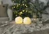 Two illuminated snowball-shaped candles are placed on a textured tablecloth in front of a blurred Christmas tree with lights and a vase with pine branches. Produktbild-Vorschau