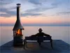 A person sits on a bench next to a fireplace on a wooden deck overlooking the ocean at sunset. Produktbild-Vorschau