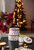 Christmas-themed cookie tins and cookies are arranged on a dark table, with a decorated Christmas tree in the background. Produktbild-Vorschau
