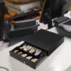 A black cash drawer is open, displaying cash and coins, on a white counter. In the background, a point-of-sale system with a monitor and keyboard is visible. Podgląd zdjęcia produktu