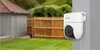 A white security camera is mounted on a gray pillar in a backyard setting, with a wooden fence and green grass in the background. Anteprima dell’immagine prodotto