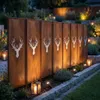 A garden scene at dusk with a metal privacy screen featuring deer head cutouts, illuminated by lanterns and garden lights. Produktbild-Vorschau