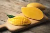 A wooden cutting board with a whole mango and a sliced mango on a wooden table. Podgląd zdjęcia produktu