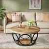 A round coffee table with a wooden top and a black metal base is placed in front of a sofa in a living room. There are decorative pillows on the sofa and a vase of flowers on the table. Produktbild-Vorschau