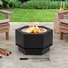 A black hexagonal fire pit with flames is situated on a paved patio, surrounded by outdoor furniture and greenery. Náhled obrázku produktu
