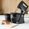 A black hand mixer with two bowls and a loaf of bread on a cutting board, set on a kitchen counter. Produktbild-Vorschau