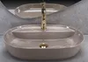 A modern beige oval countertop sink with a gold faucet, set against a textured gray background and a reflection in a mirror. Náhled obrázku produktu