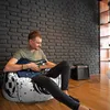 A man is sitting on a bean bag chair and playing a guitar in a room with a brick wall. Voir les images du produit