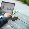 A person is using a laptop and a tablet outdoors, with a power bank attached to a smartphone on the table. Produktbild-Vorschau
