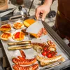 A person is using a spatula to flip bacon on a griddle with various food items. Podgląd zdjęcia produktu