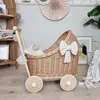 A wicker doll stroller with a bow and blanket is placed on a wooden floor in a room with shelves and wall decorations. Produktbild-Vorschau