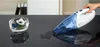 A hand is holding a blue and white handheld vacuum cleaner over a dark, reflective surface, with a small glass bowl containing flowers and water to the left. A spill of liquid is also visible on the surface. Podgląd zdjęcia produktu