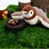 Braun-weißes Plüschvogelspielzeug mit gelbem Schnabel, neben einem kleinen Nest mit zwei Eicheln, auf grünem Moos Náhľad obrázku produktu